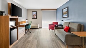 Une chambre d’hôtel moderne dotée d’une kitchenette, d’un téléviseur à écran plat, d’un canapé rouge, d’une table en bois et d’une grande fenêtre offrant une vue sur le paysage urbain.