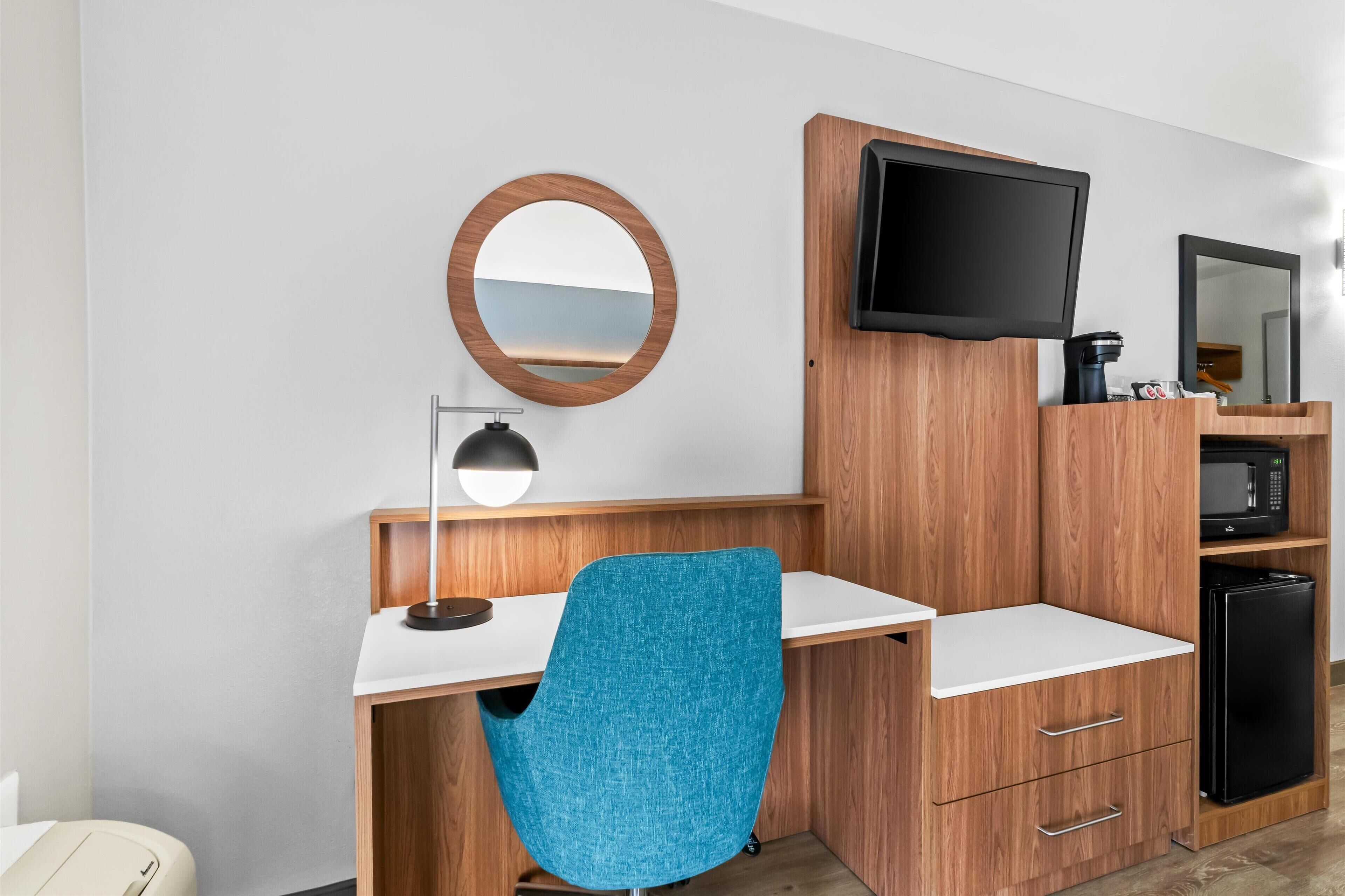 A modern hotel room with a wooden desk, a blue chair, a flat-screen TV, a coffee machine, a microwave, and a refrigerator.
