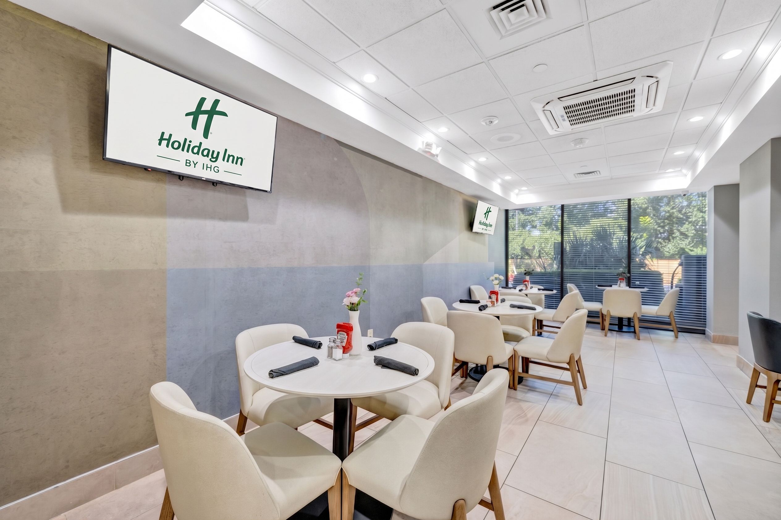 Ein Holiday Inn Hotelzimmer mit einem großen Bildschirm, der den Namen des Hotels zeigt, mehreren Sitzbereichen mit Tischen und Stühlen sowie einem Blick ins Freie durch große Fenster.