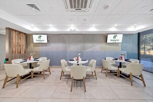 A conference room with four tables and chairs, two large screens displaying the Holiday Inn logo, and a large window with blinds.