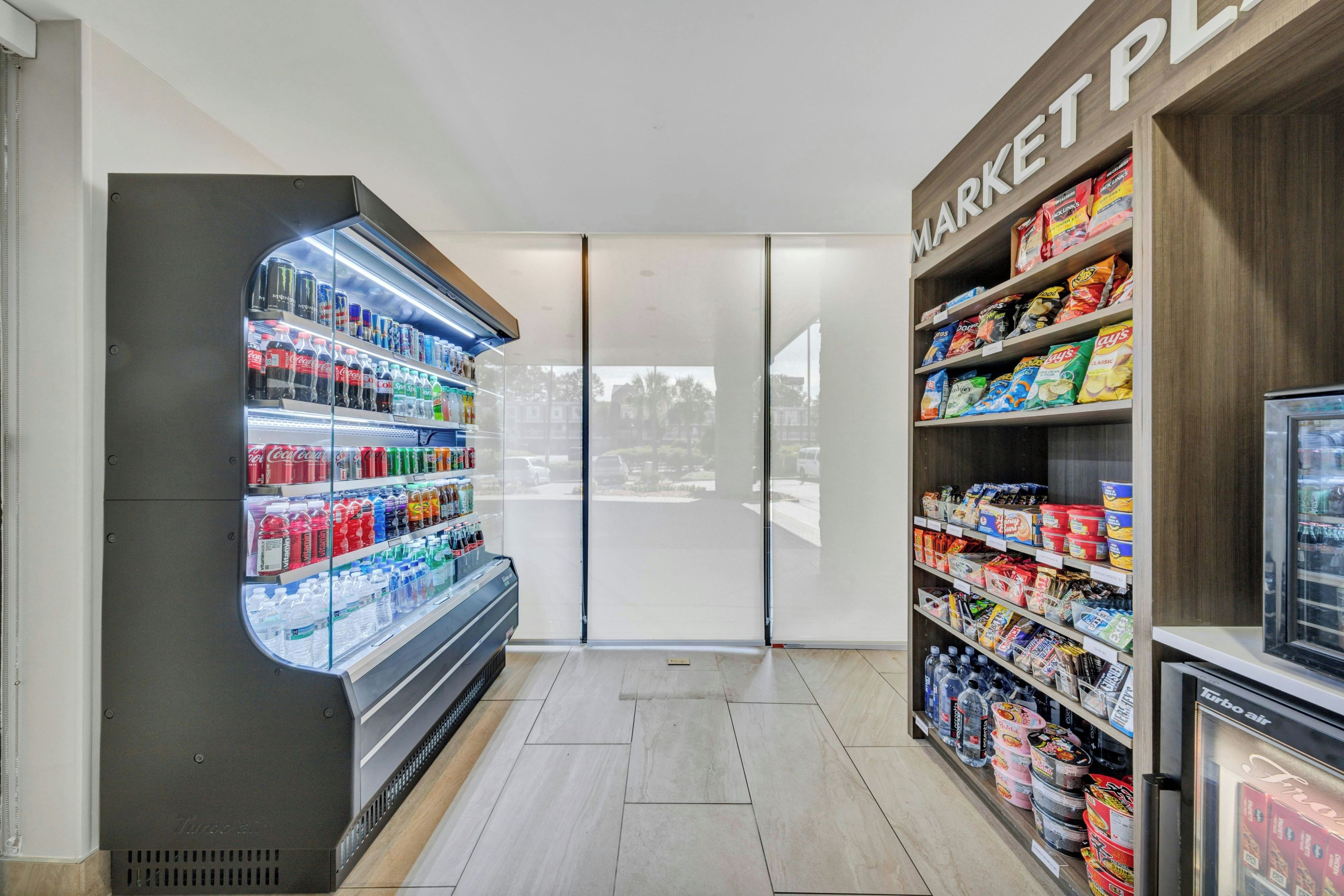 Interno di un negozio di alimentari con un banco frigo per bevande, scaffali di snack e una buona visuale sull'esterno.