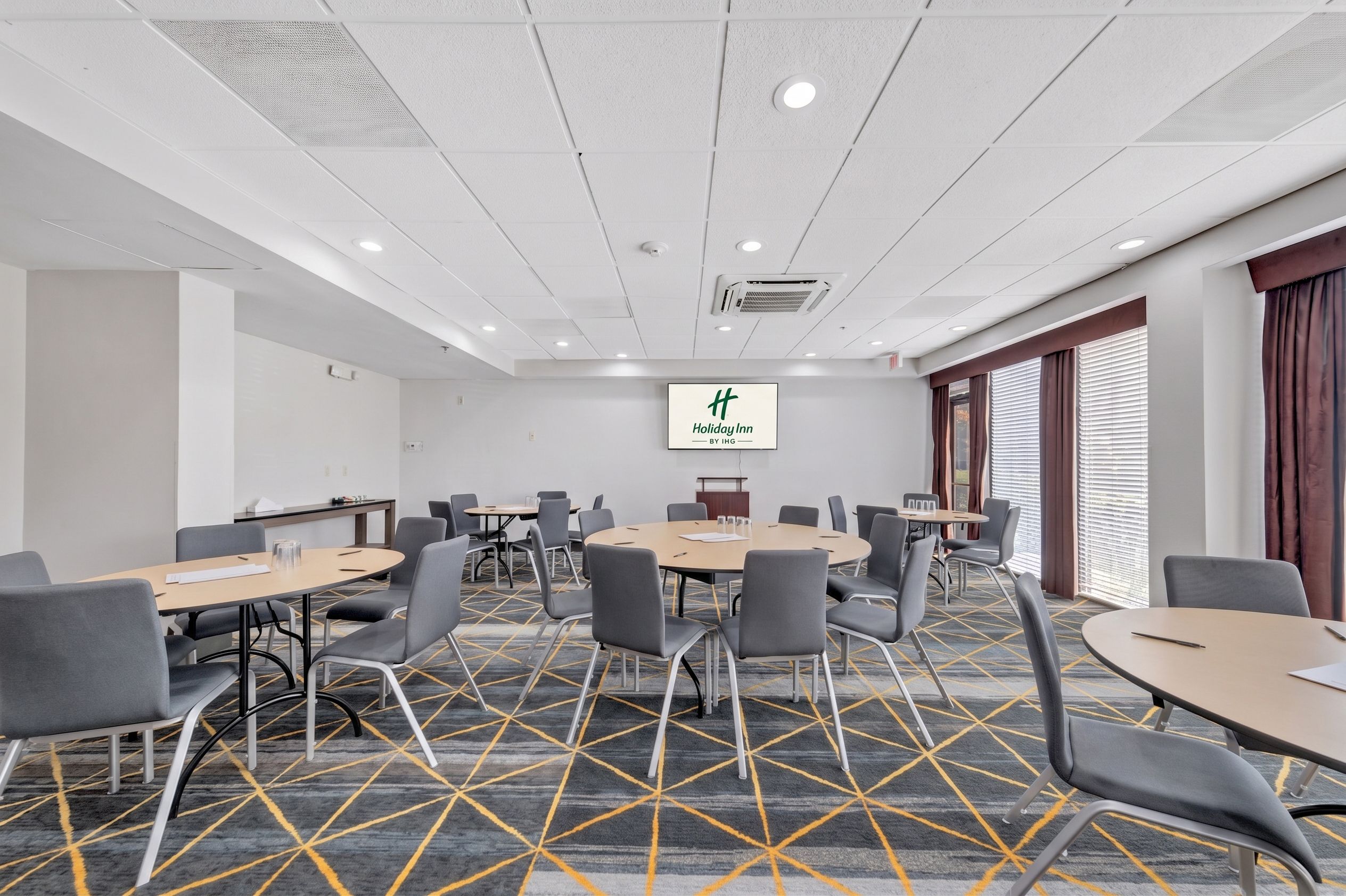 A conference room with a large screen displaying 'Holiday Inn by IHG' logo, multiple tables with chairs, and a patterned carpet.
