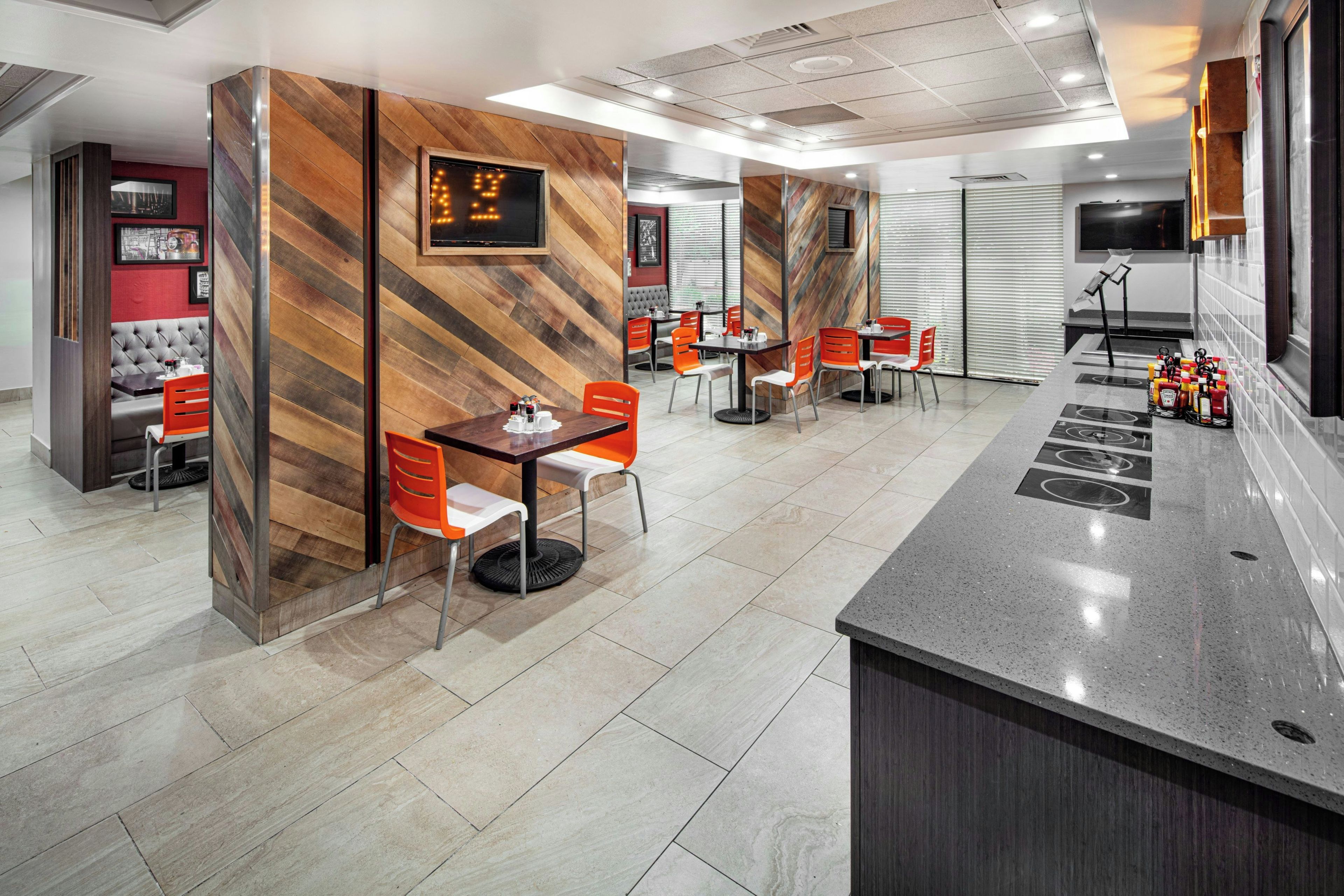 Ein modernes Restaurant mit Holzwandverkleidung, orangefarbenen Stühlen und einer zentralen Theke mit Gewürzspendern.