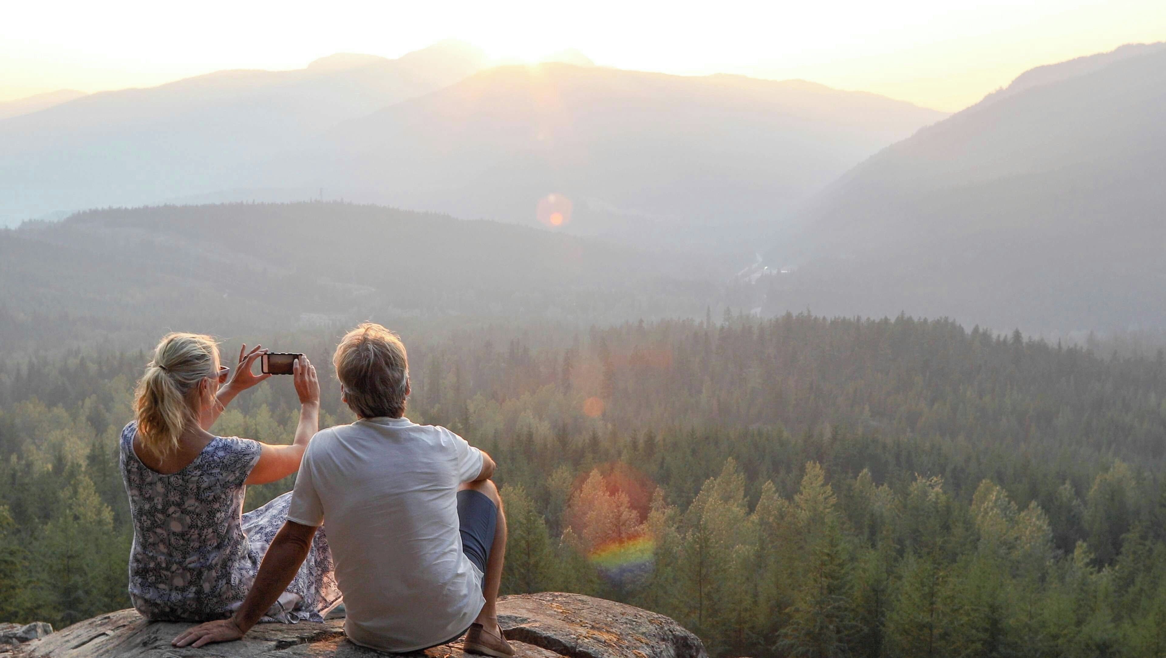 Zwei Personen sitzen auf einem Felsen, im Hintergrund ein Wald und Berge.