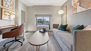 A modern hotel room with a wooden desk, a brown leather chair, a grey sofa with green cushions, a round wooden table, and a large window with a view of buildings. - Holiday Inn Atlanta-Northlake by IHG (Atlanta)