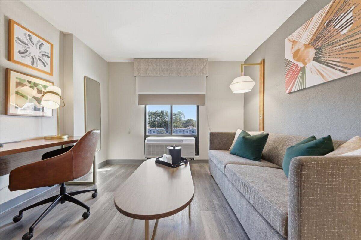 Une chambre d’hôtel moderne avec un bureau en bois, un fauteuil en cuir marron, un canapé gris avec des coussins verts, une table ronde en bois et une grande fenêtre donnant sur des immeubles.