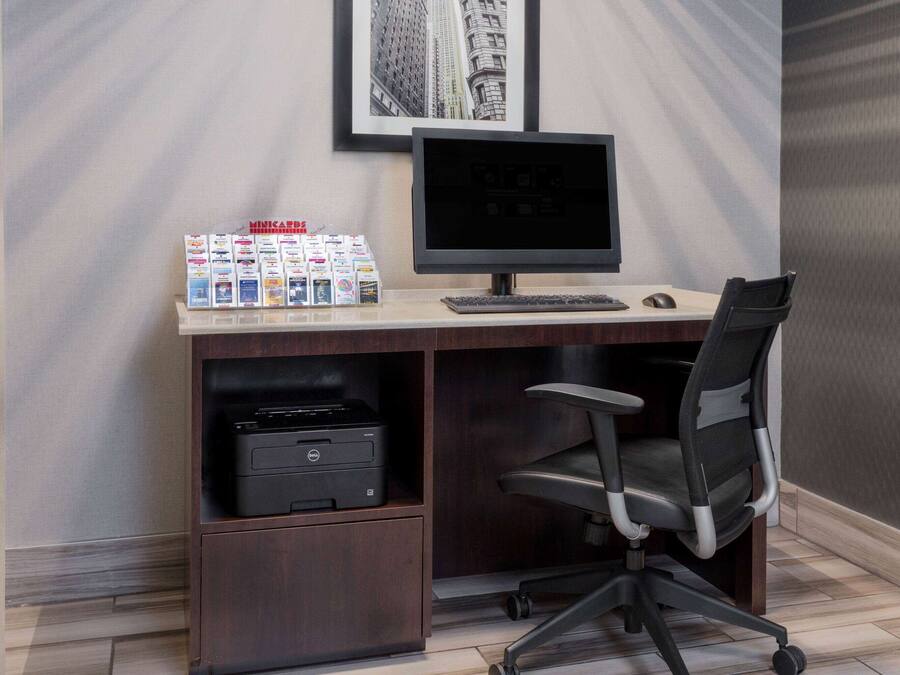 A modern office desk with a computer, keyboard, mouse, and office chair. There are magazines on the desk, and a printer is visible in the lower shelf.