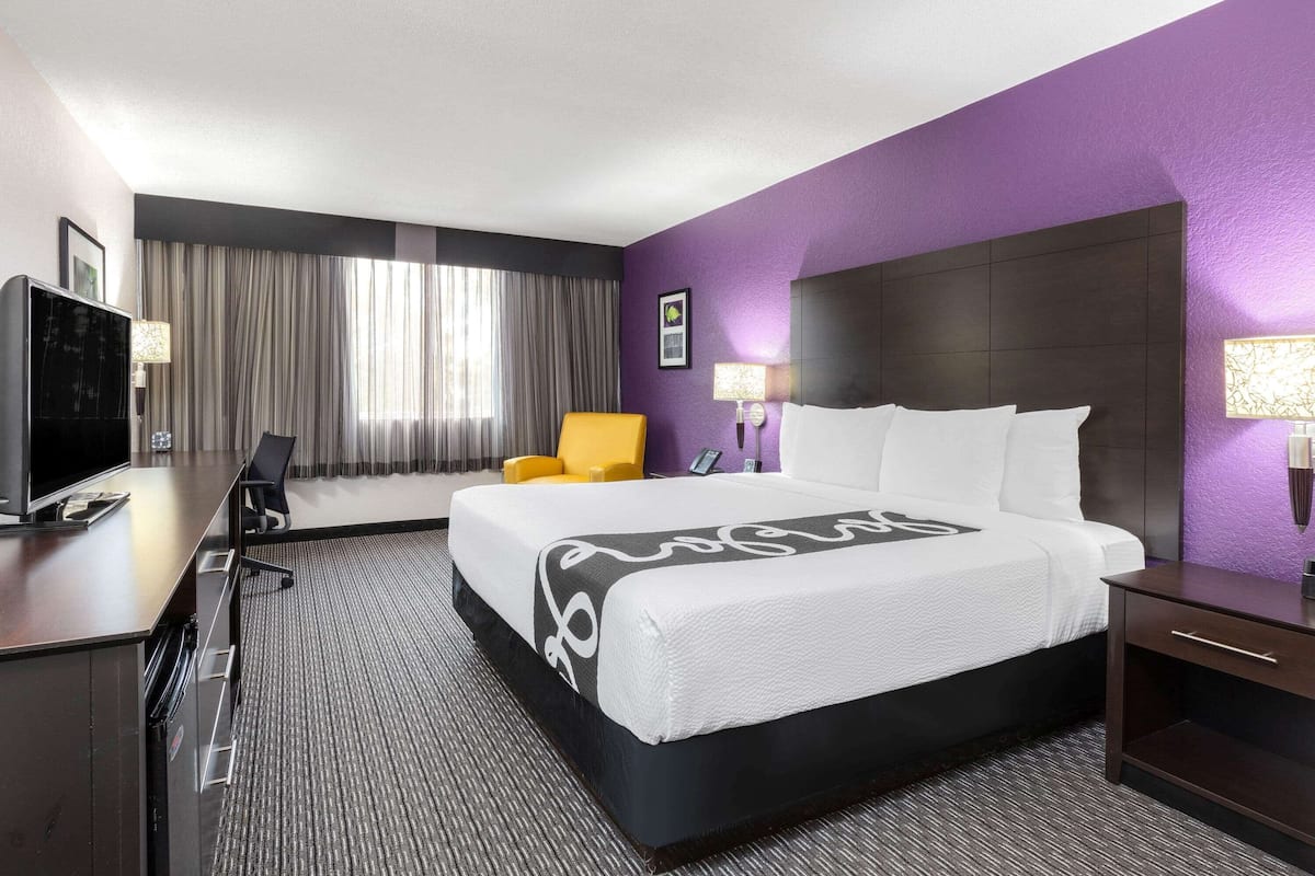 Una habitación de hotel con una cama grande, un escritorio con televisor, una silla y un ventanal con cortinas.