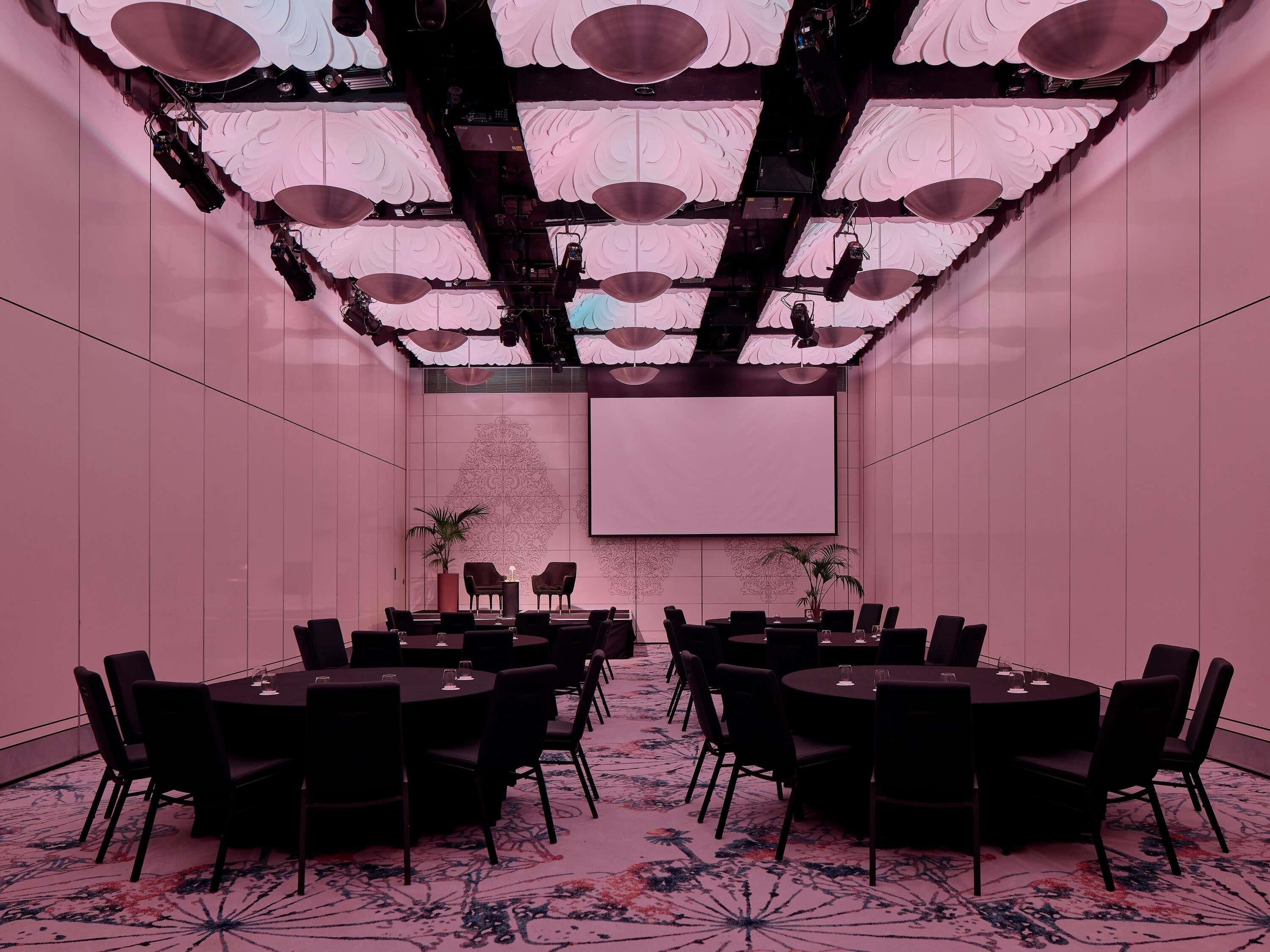 A conference room with multiple tables and chairs, a projector screen, and a decorative ceiling.