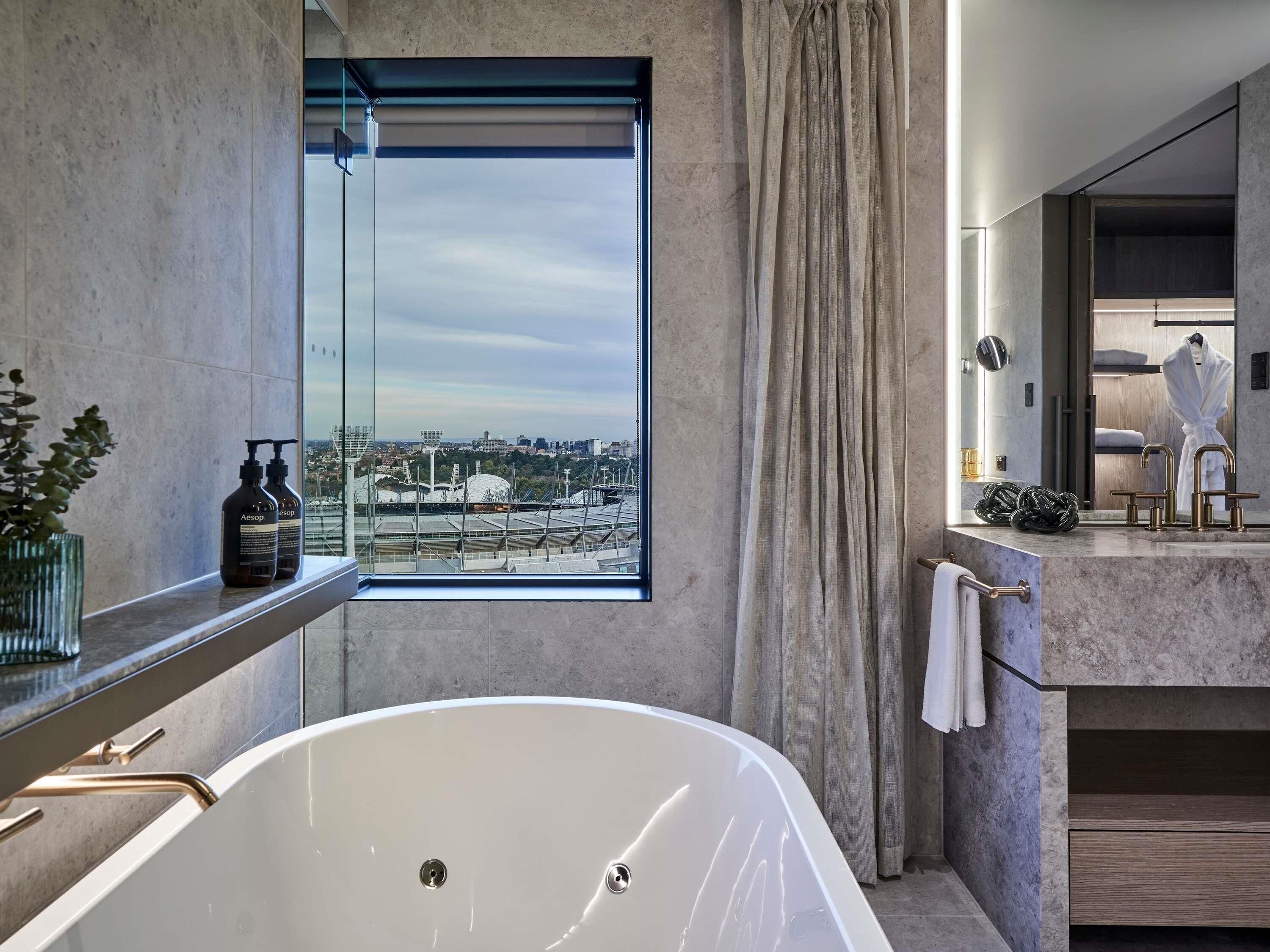 A modern bathroom with a large bathtub, a window with a city view, a vanity with a mirror, and a robe hanging on the wall.