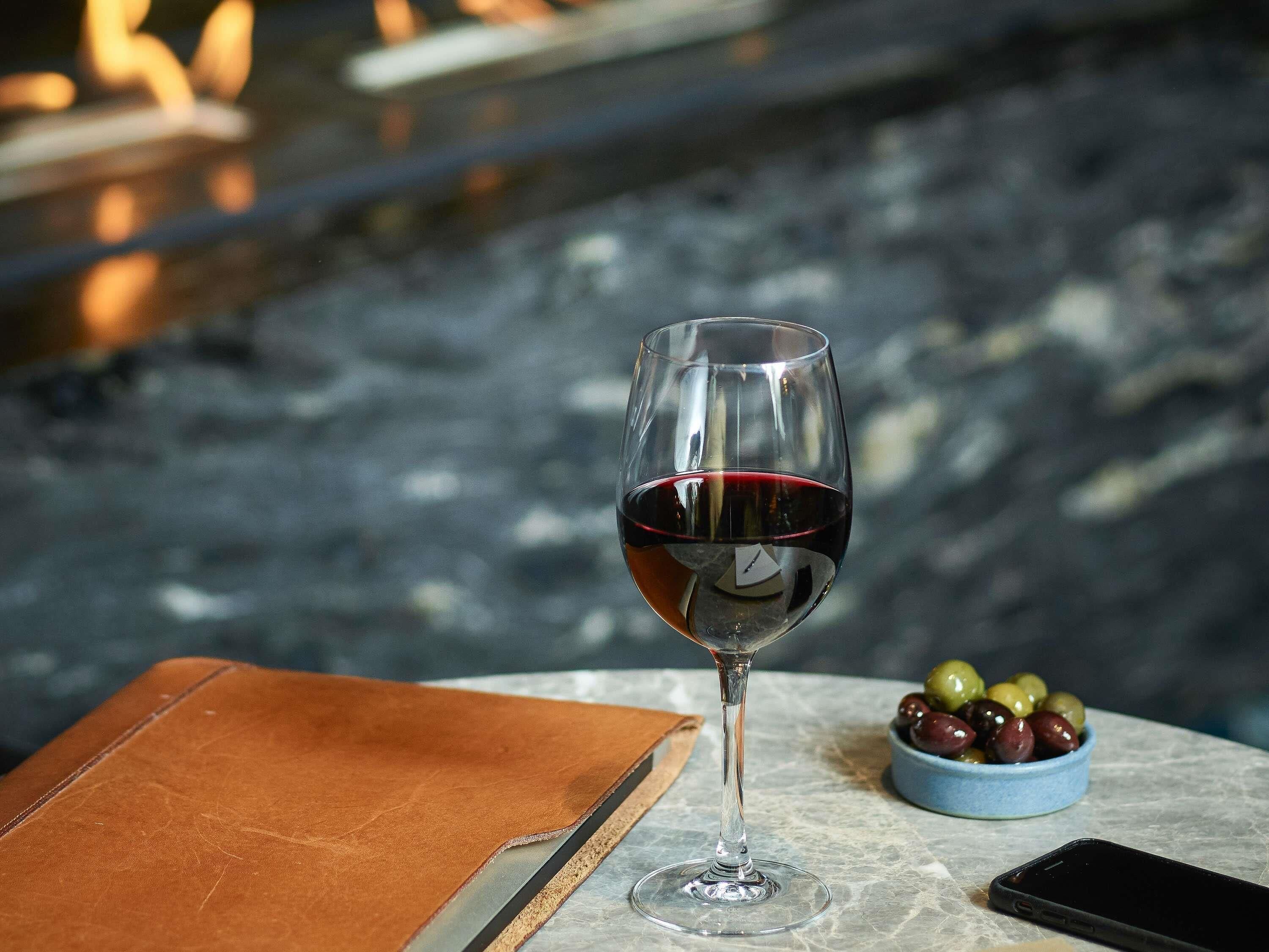 Een wijnglas met rode wijn op een marmeren tafel, een leren notitieboek, een smartphone en een schaal olijven.