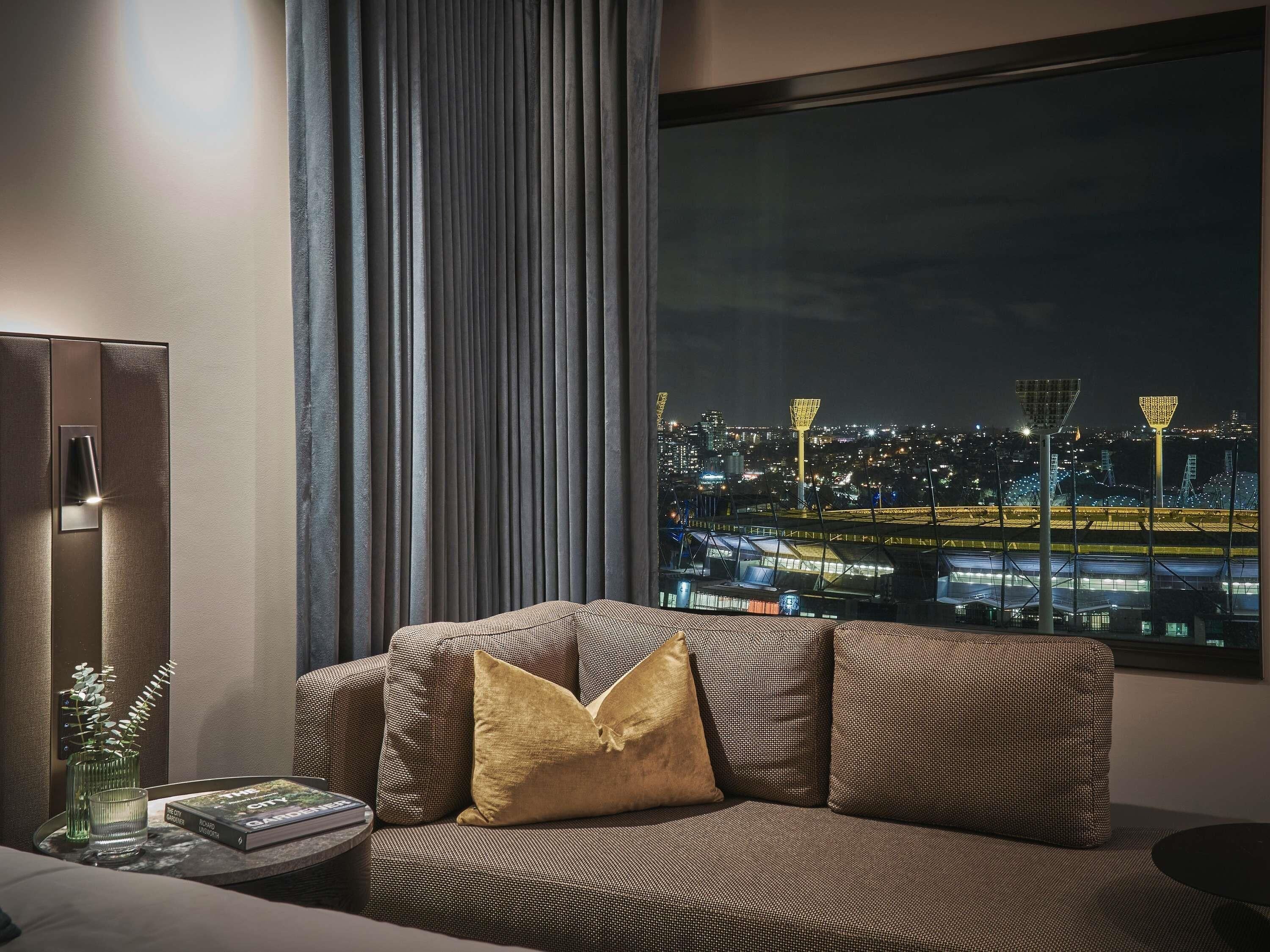 Une chambre d’hôtel moderne dotée d’une grande fenêtre offrant une vue sur la ville la nuit, d’un canapé avec des coussins, d’une table d’appoint avec un livre et un verre, et d’une lampe.