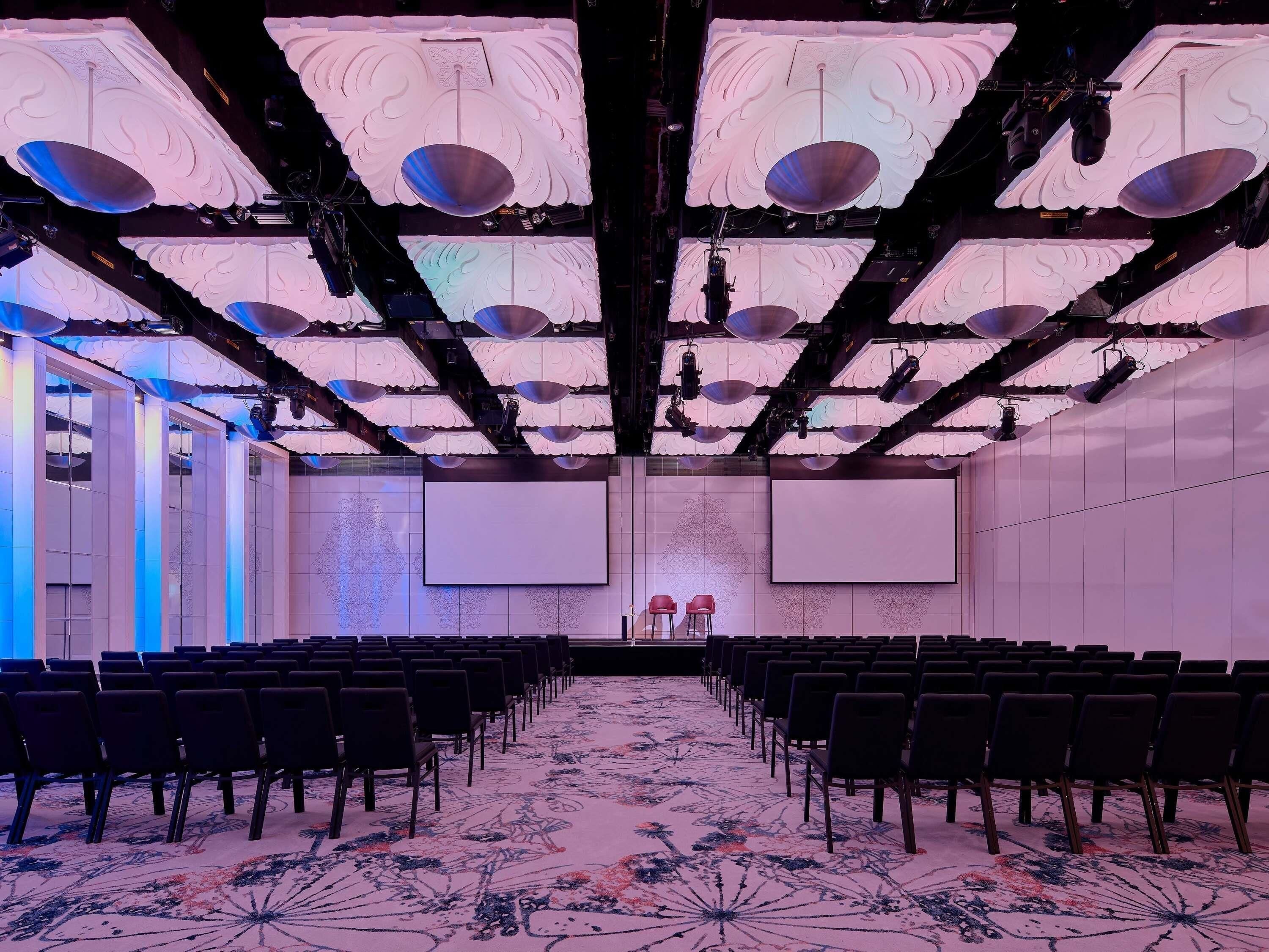 Un salón de conferencias con filas de sillas, una pantalla grande e iluminación decorativa en el techo.