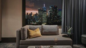 A modern living room with a cityscape view, a sofa with patterned cushions, a glass of red wine, and a small table with a vase. - Pullman East Melbourne (East Melbourne)