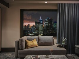 A modern living room with a cityscape view, a sofa, a small table with a glass of red wine, and a vase with plants.