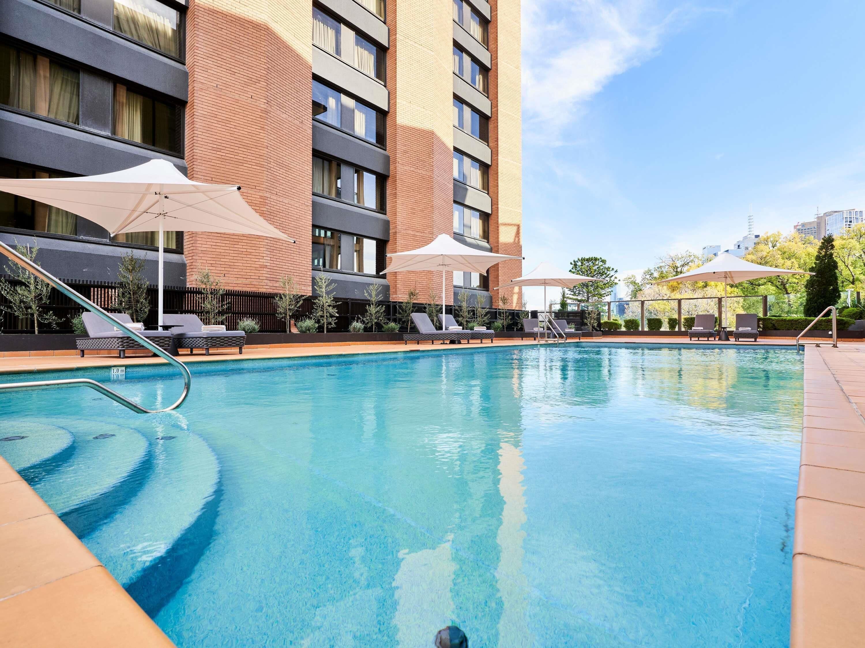Una piscina con sillas de descanso y sombrillas, rodeada por un edificio de varios pisos y ventanas.