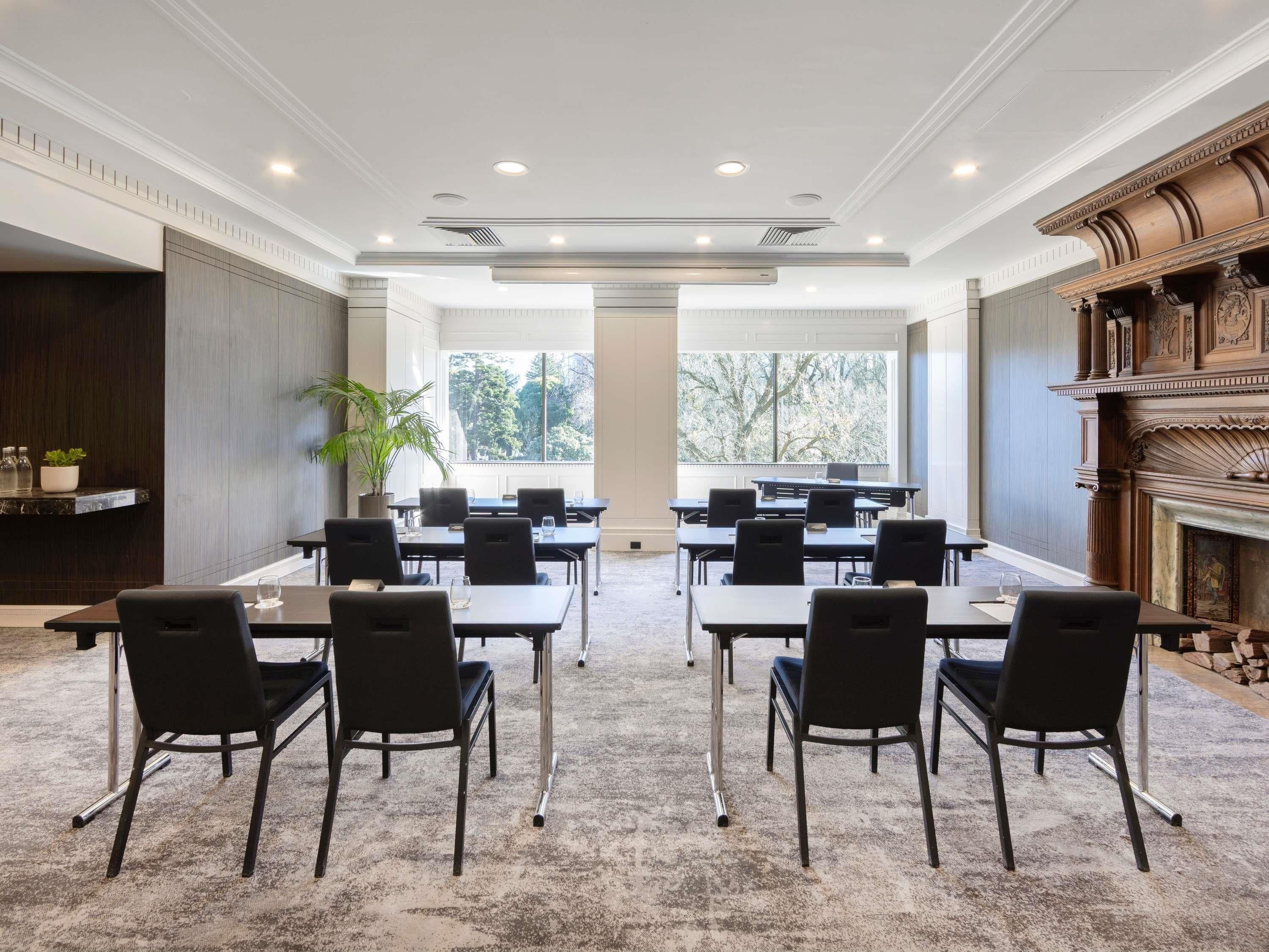 Un salón de conferencias con varias mesas y sillas, amplias ventanas que dan a vistas de árboles y una chimenea con elaborados detalles de madera.