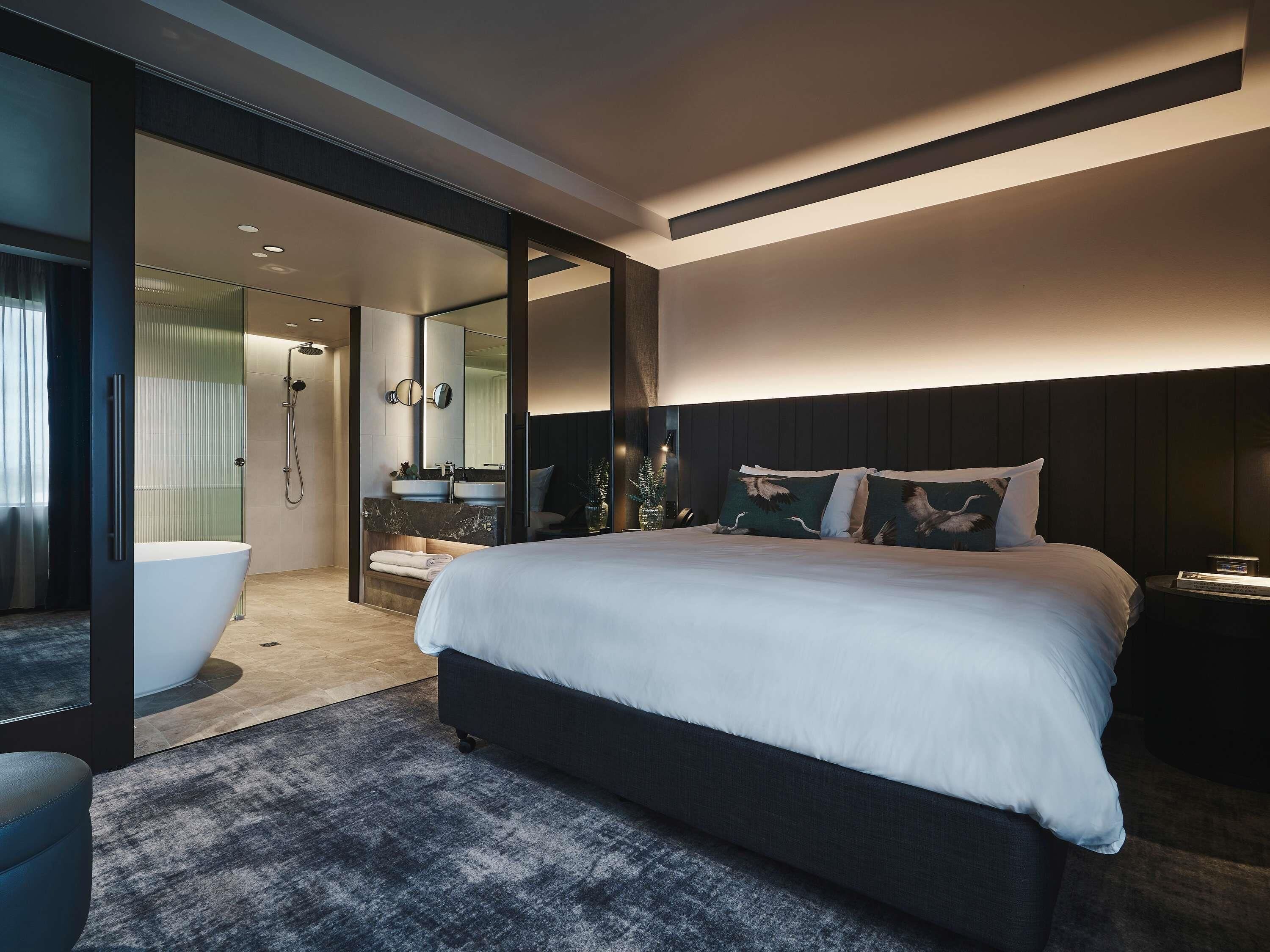 Une chambre moderne avec un grand lit, une douche à l’italienne, une salle de bain avec un lavabo et un miroir, et une tête de lit de couleur sombre.