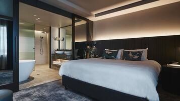Une chambre moderne avec un grand lit, une douche à l’italienne, une salle de bain avec un lavabo et un miroir, et une tête de lit de couleur sombre.