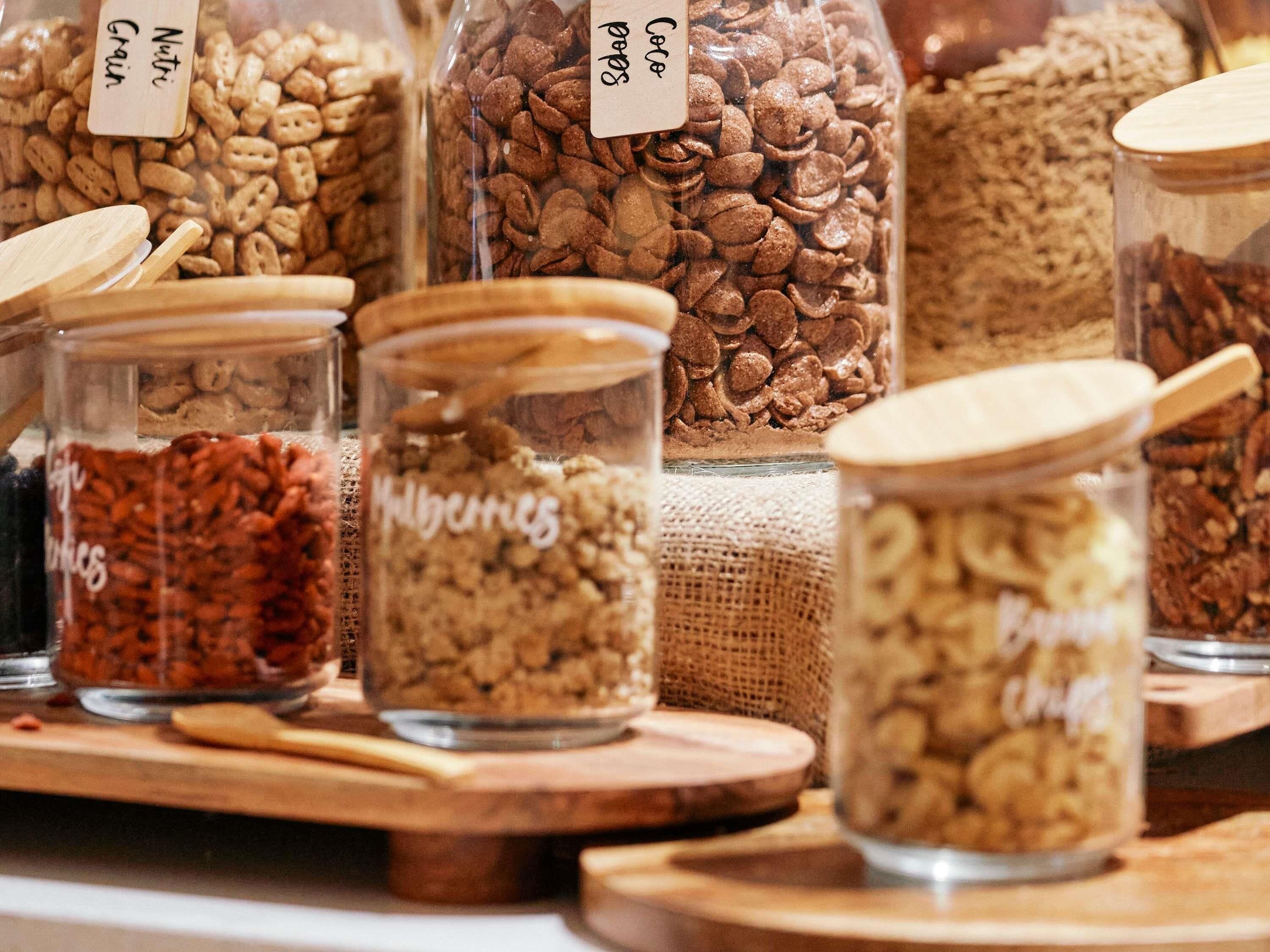A variety of food items in glass jars with wooden lids, labeled with names like 'Nutri Grain' and 'Cocoa.'