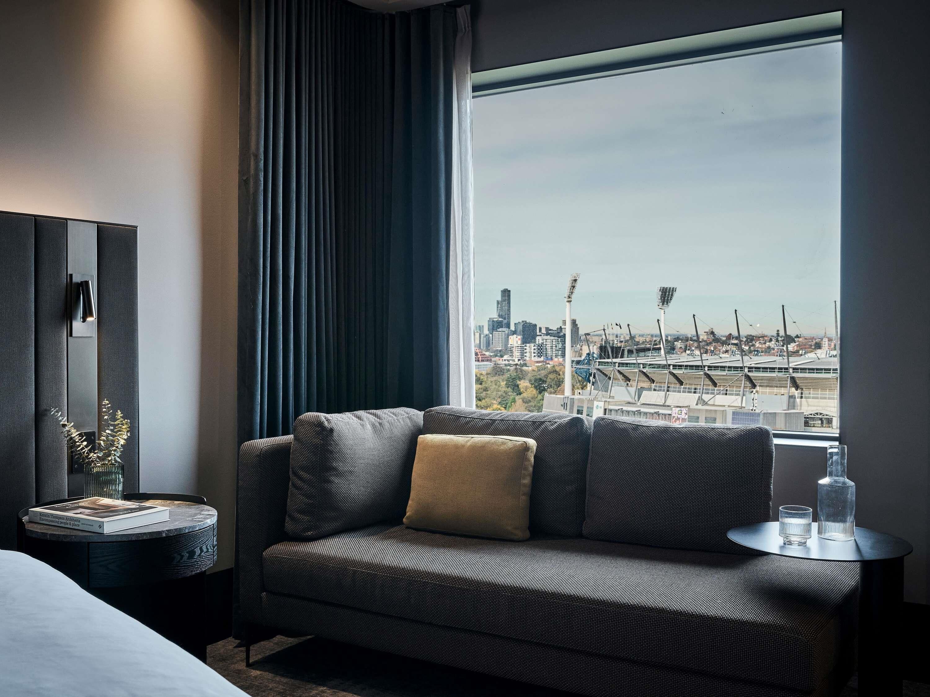 Ein modernes Hotelzimmer mit einer Couch, einem kleinen Beistelltisch, einem Spiegel und einem großen Fenster mit Blick auf die Stadt.