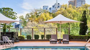 A rooftop pool with lounge chairs and umbrellas, surrounded by trees and city buildings. - Pullman East Melbourne (East Melbourne)