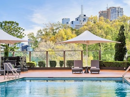 A rooftop pool with lounge chairs and umbrellas, surrounded by trees and city buildings.