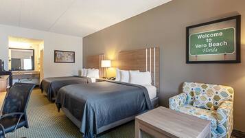 A hotel room with two beds, a desk, a chair, a small table, and a framed welcome sign.
