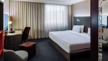 A hotel room with a large bed, a desk with a chair, a small ottoman, and a television.