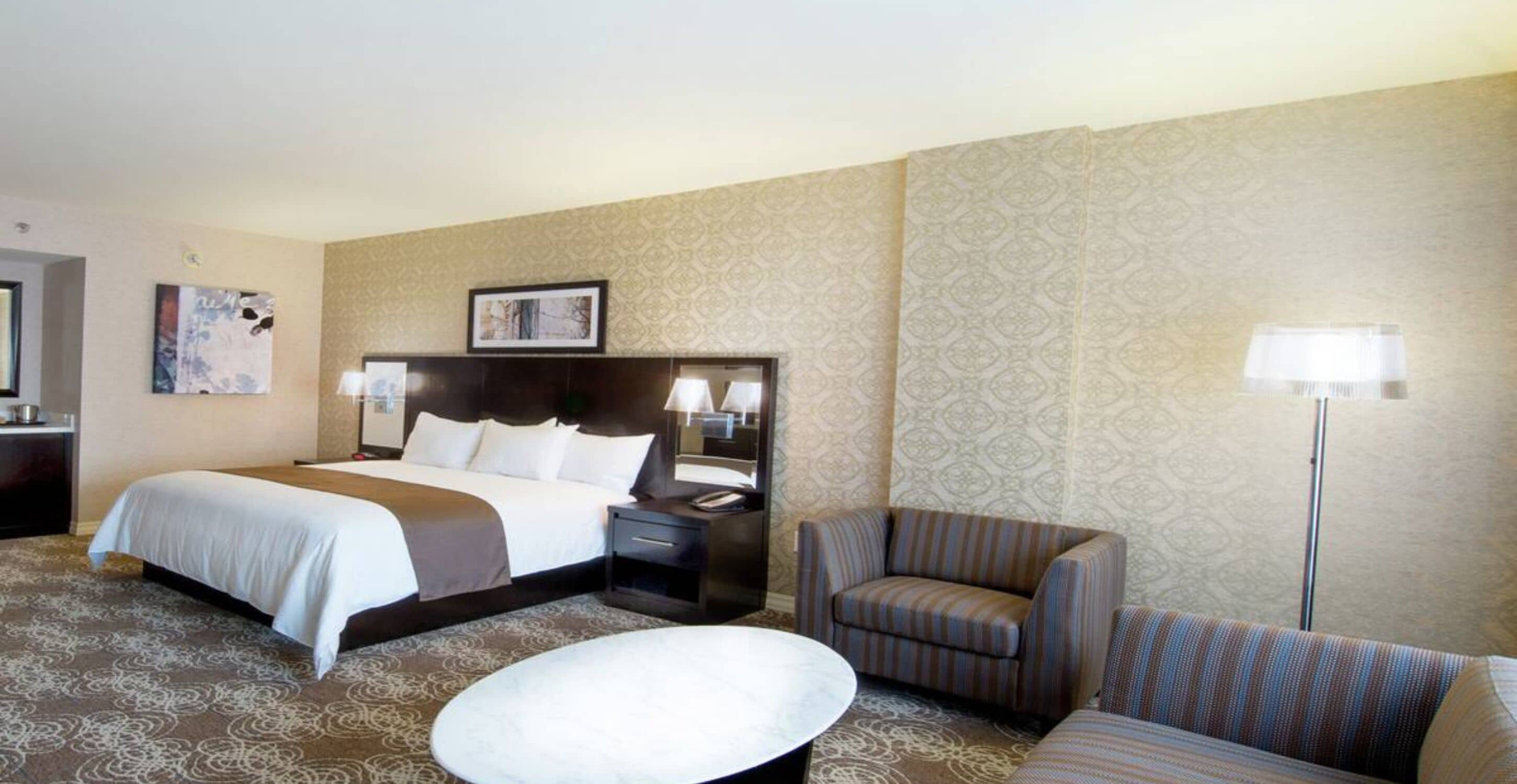 Ein Hotelzimmer mit einem großen Bett, einer Sitzecke mit einem Sessel, einem kleinen Tisch und einem Nachttisch mit Lampe.