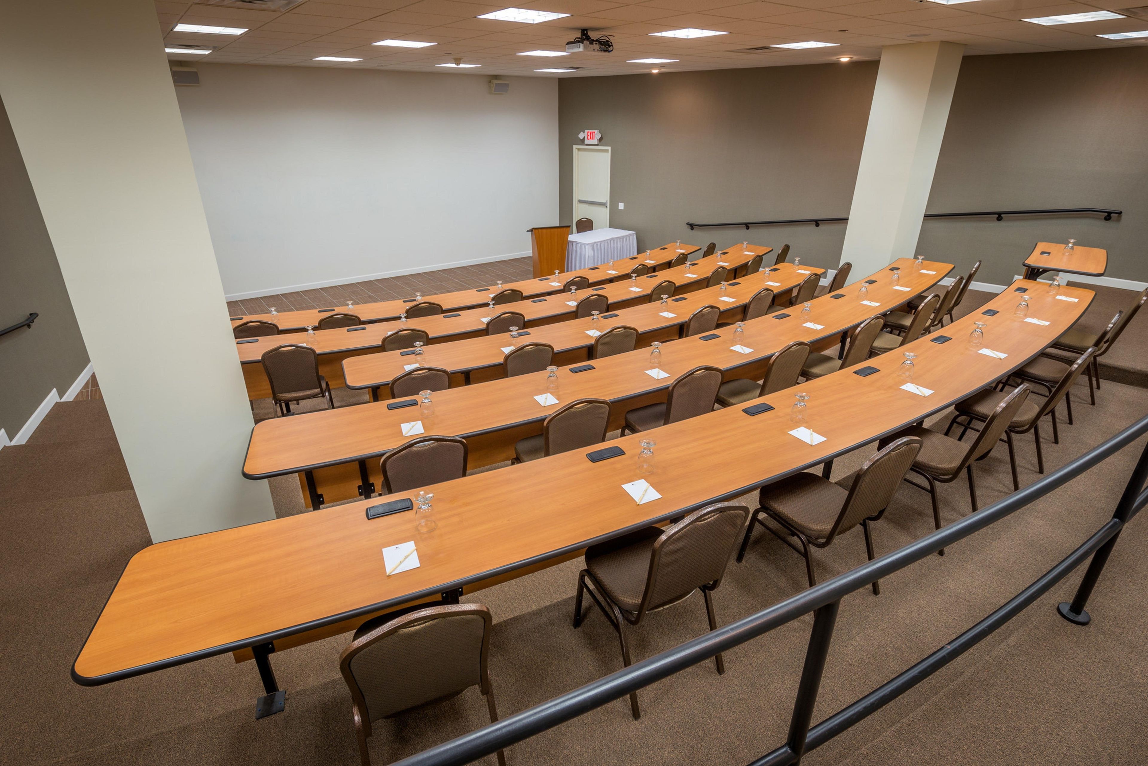 A conference room with multiple rows of tables and chairs, a podium, and an exit sign.