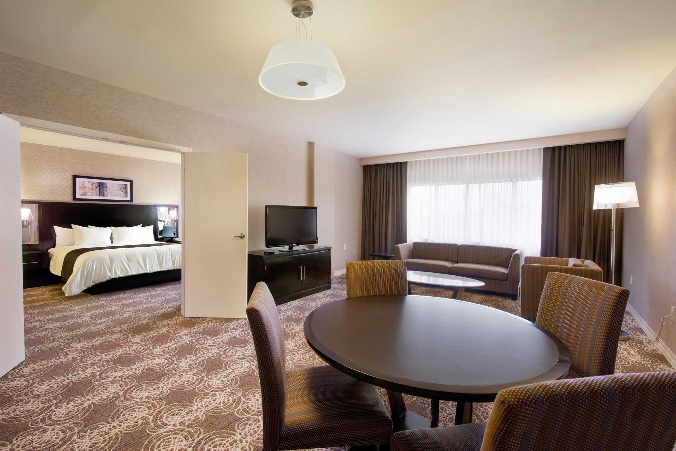 A hotel room with a bed, a TV, a seating area with a table and chairs, and a large window with curtains.