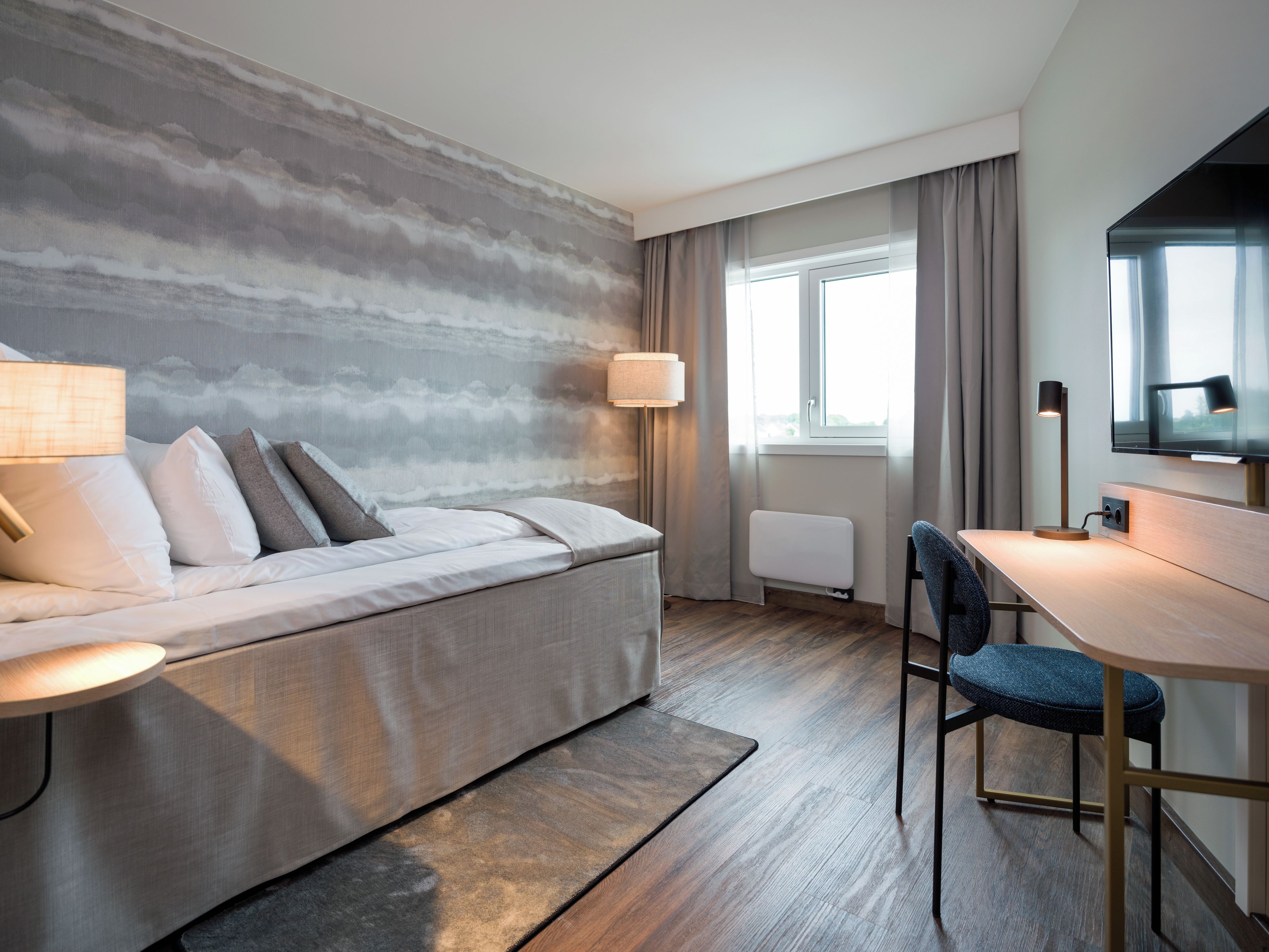A modern hotel room with a sofa, a desk, a chair, a lamp, a radiator, and a TV.