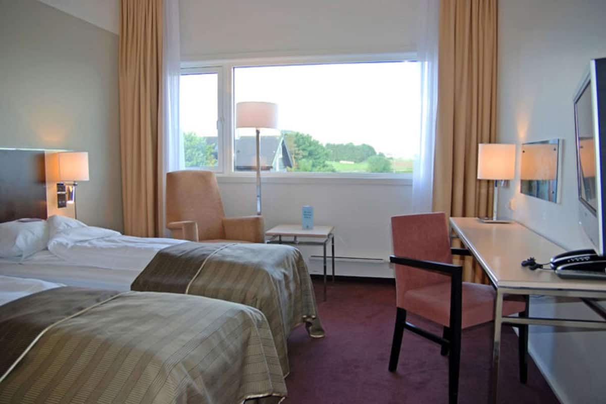 Habitación de hotel con dos camas, un escritorio, una silla y una ventana con vistas a zonas verdes.