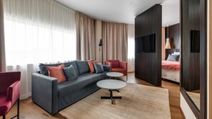 Un salon moderne avec un canapé gris, un fauteuil rouge et une petite table ronde. On aperçoit un lit recouvert d’une couverture rouge à l’arrière-plan.