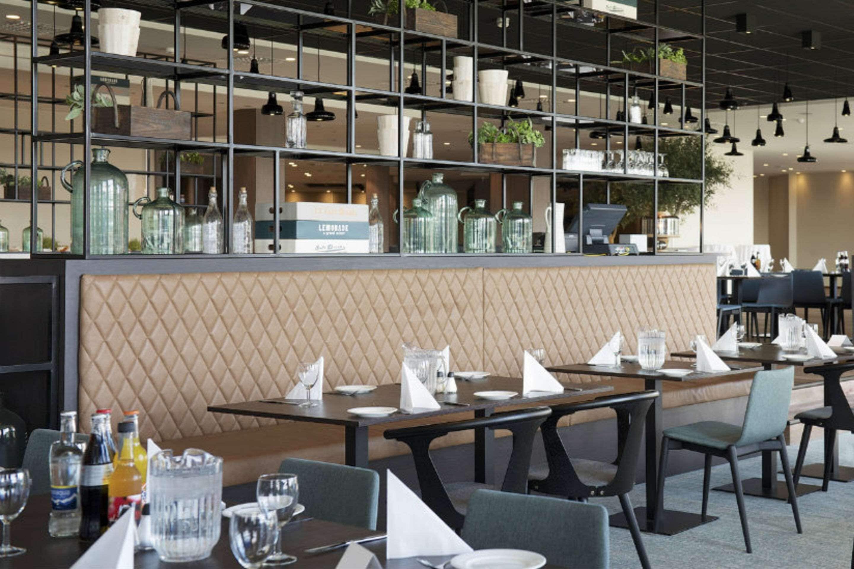 A restaurant with a modern interior, featuring a long counter with neatly arranged tables and chairs, a decorative wall with a geometric pattern, and shelves with glassware and plants.