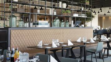 A restaurant with a modern interior, featuring a long counter with neatly arranged tables and chairs, a decorative wall with a geometric pattern, and shelves with glassware and plants.