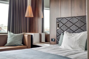 Een moderne hotelkamer met een bed, een zithoek met een bank en kussens, een hanglamp en een bedieningspaneel aan de muur.