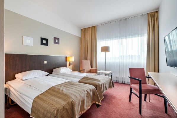 A hotel room with two beds, a desk, a chair, a TV, and a window with curtains.