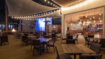 Un espacio exterior para comer con mesas y sillas, una chimenea y luces colgantes.