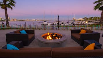 A fire pit surrounded by wicker furniture with cushions, overlooking a marina with boats and a city skyline in the background.
