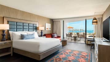 A hotel room with a large bed, a desk with a TV, a balcony with outdoor seating, and a view of the sea.