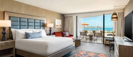 A hotel room with a large bed, a desk with a TV, a balcony with outdoor seating, and a view of the sea.