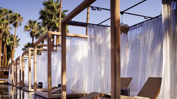 Zona junto a la piscina con tumbonas, cortinas blancas y palmeras al fondo.