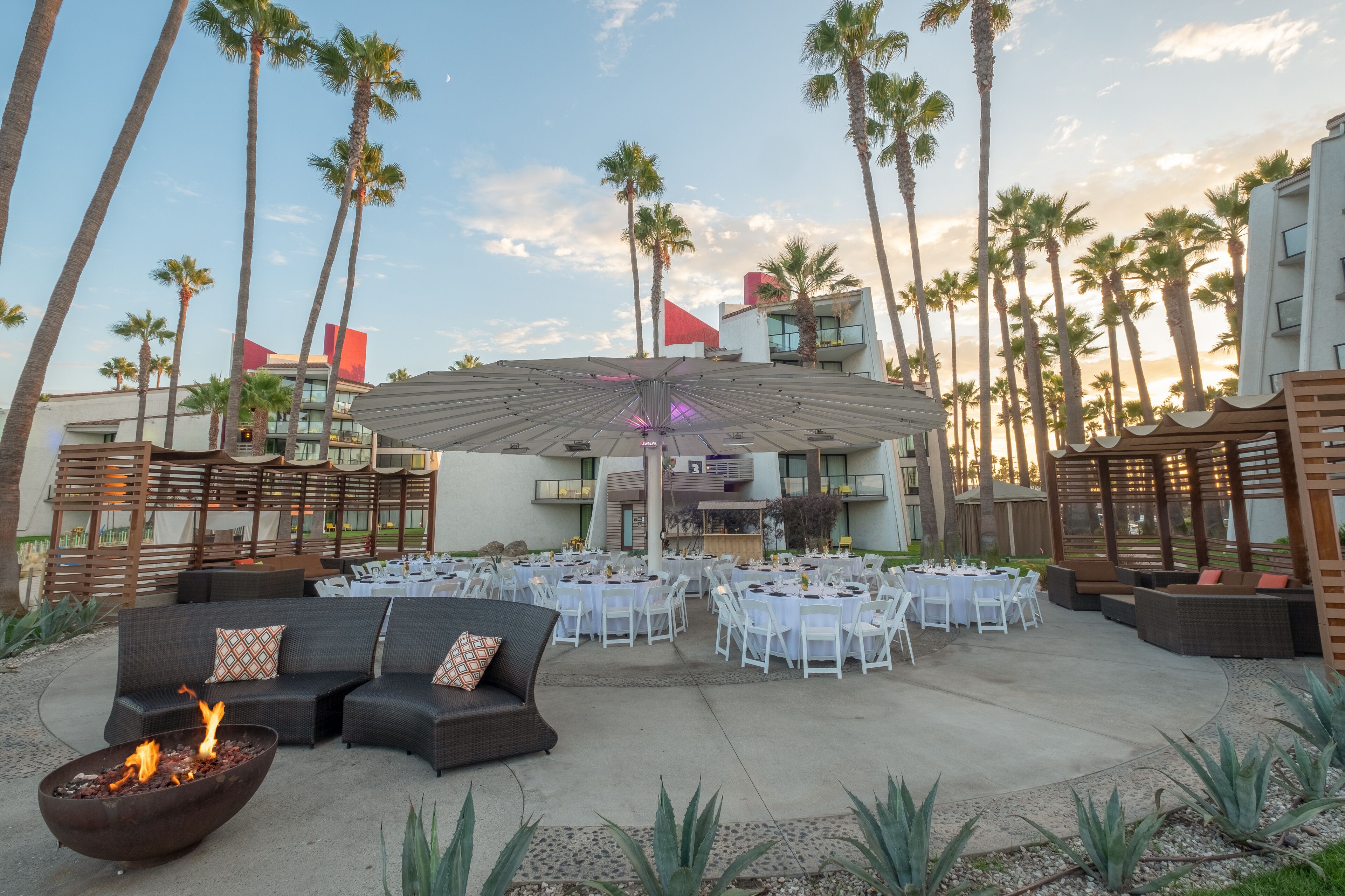 An outdoor patio with round tables and chairs, a fire pit, and a large umbrella.