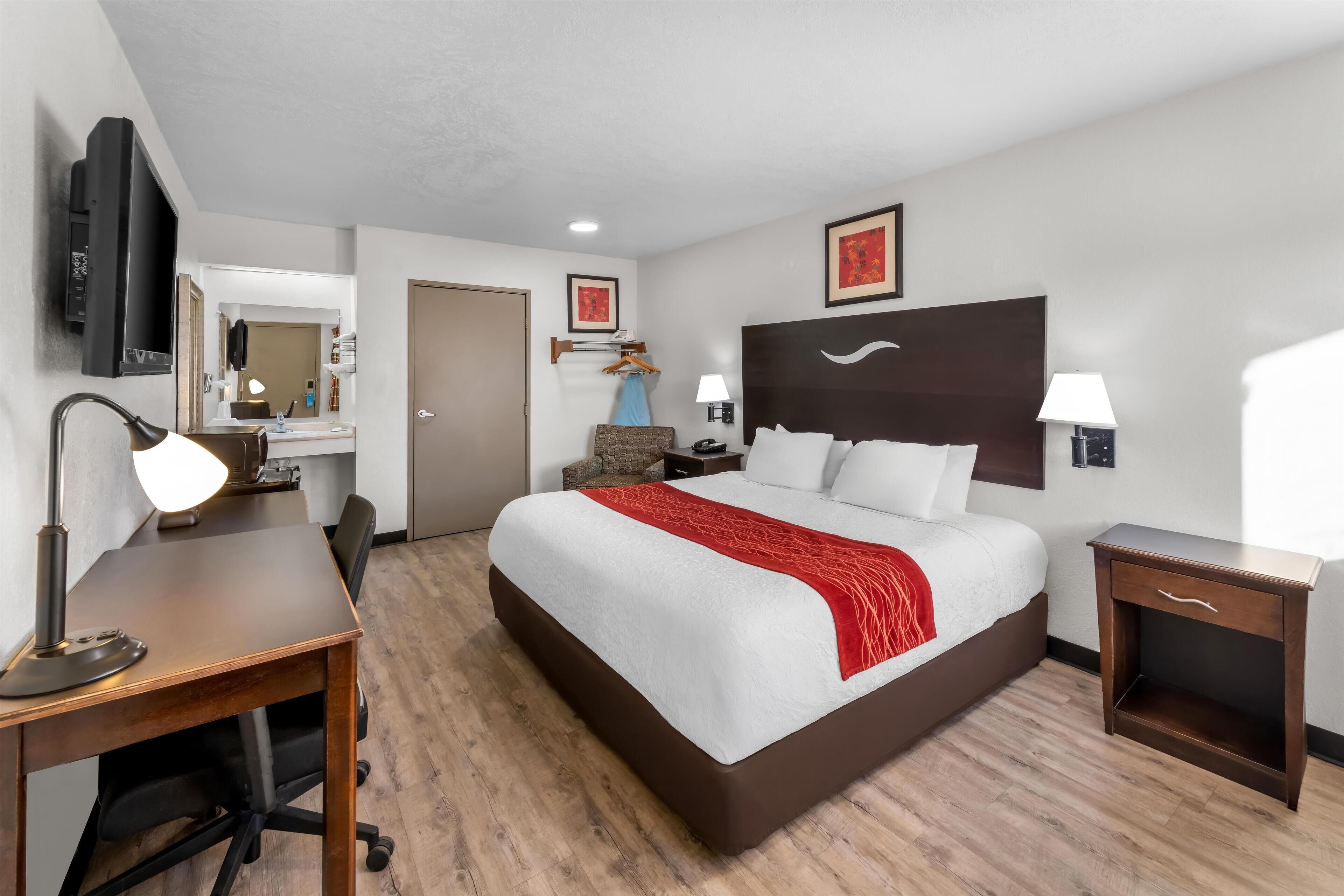 A hotel room with a large bed, a desk with a chair, a TV, a bedside table with a lamp, and a mirror.