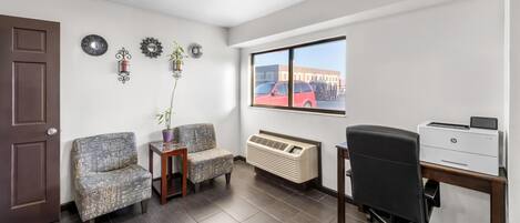 A room with a brown door, two patterned chairs, a small table with a plant, a window with a red car view, and a desk with a printer.