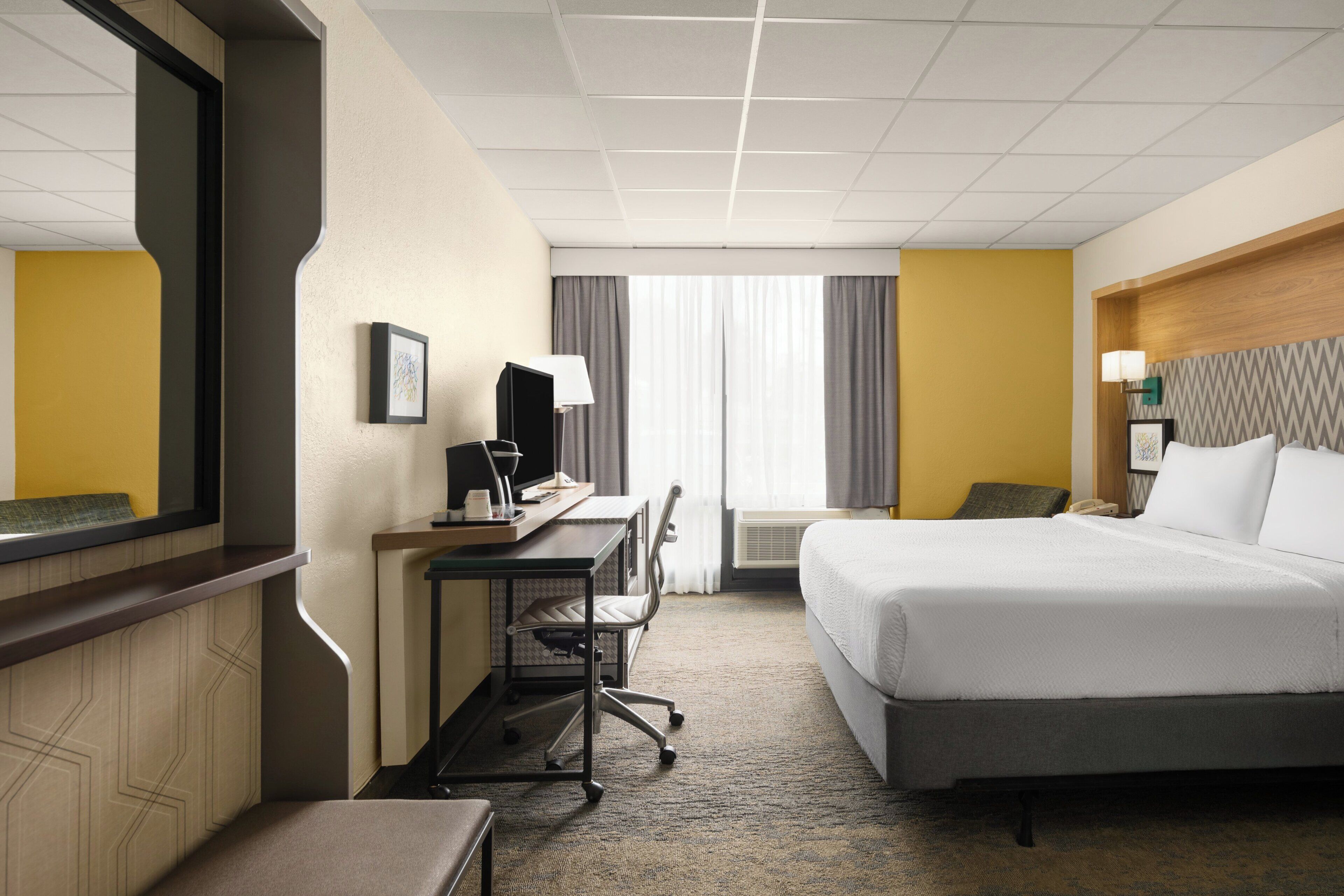 A hotel room with a bed, desk, chair, and a large mirror.