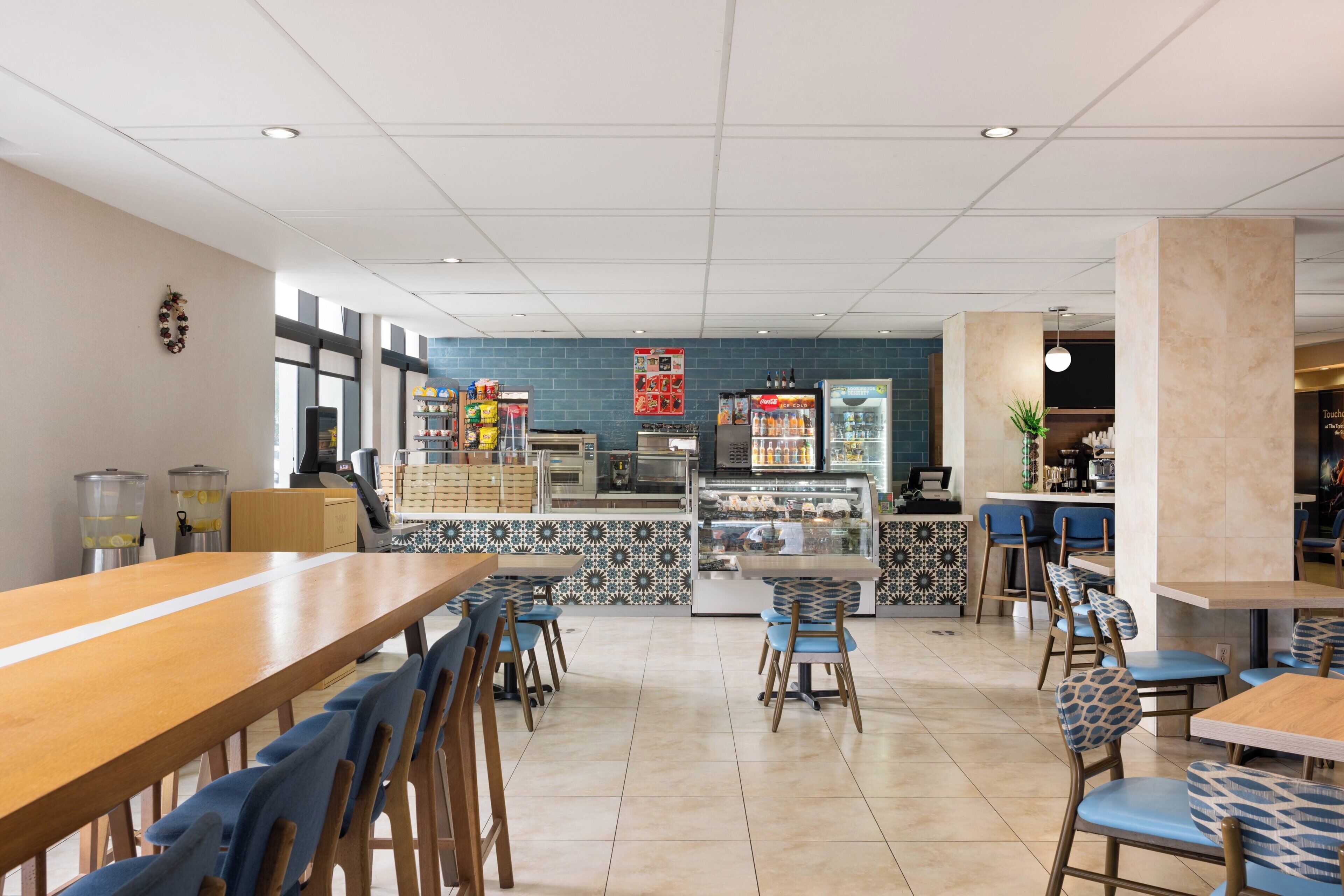 L’intérieur d’un restaurant présente une longue table en bois, des chaises à motifs bleus, un comptoir où sont exposés des plats, et un mur recouvert de carreaux.