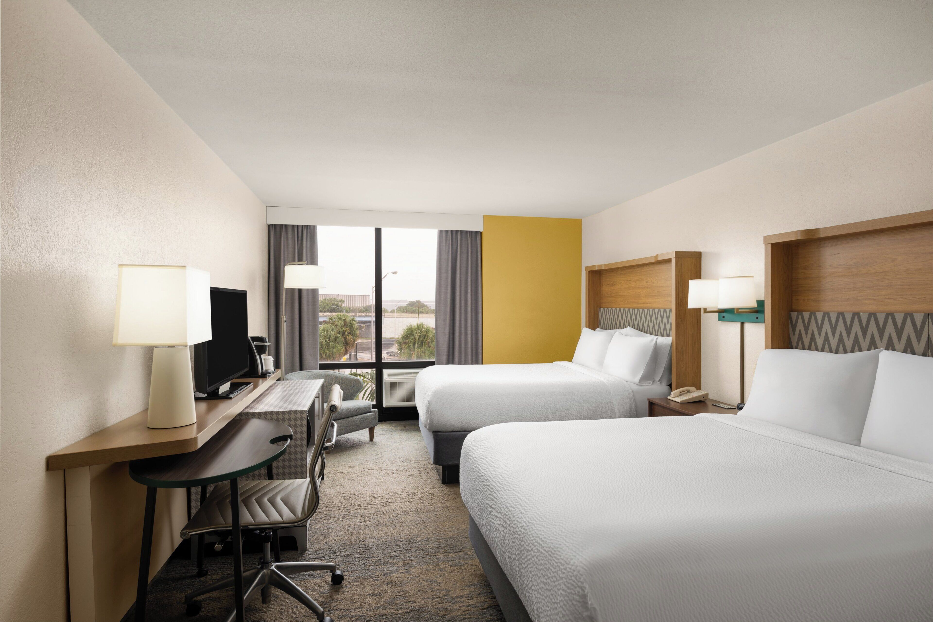 Habitación de hotel con dos camas, un escritorio con computadora, una silla y un ventanal con cortinas.